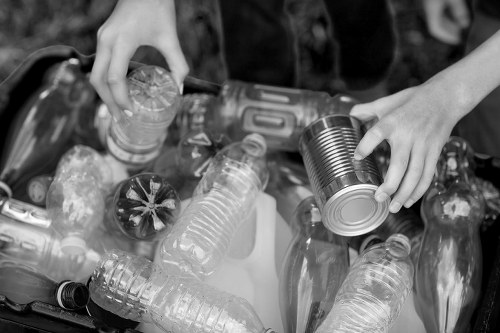 A pair of hands are shown sorting through a collection of recyclable waste, specifically plastics and metal cans, in an outdoor environment. The person is holding a metal tin can in the right hand, while the left hand is reaching for a plastic bottle among other discarded bottles and containers. The plastic bottles vary in shape and size, with some appearing crushed or partially flattened, made of clear or semi-transparent plastic materials. The cans and bottles are loosely piled together on a surface, possibly in a recycling or rubbish collection context associated with waste removal services. The environment suggests an outdoor setting, with blurred background foliage or natural surroundings. The lighting appears natural, highlighting the textures of the plastic and metal objects. This detailed scene is relevant to rubbish removal and recycling operations in Redbridge or nearby areas, where Redbridge House Clearance may assist with waste collection and disposal.