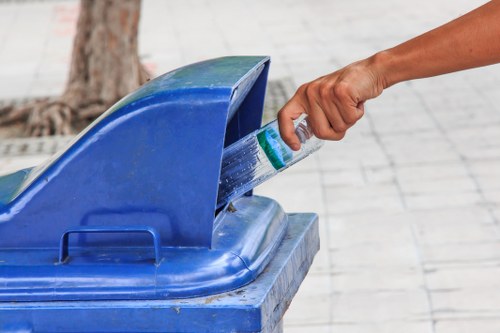 A person's hand is seen placing a plastic bottle into the open top of a blue recycling bin located outdoors on a paved surface. The bin appears to be made of sturdy plastic with a slightly textured finish, and the lid is tilted back to reveal the opening. In the background, there is a tree trunk and some blurred pavement, suggesting a residential or community area. The scene reflects the act of waste recycling, which aligns with rubbish removal and disposal services offered by companies such as Redbridge House Clearance, based in the Redbridge area. The lighting is natural, indicating daytime, and the overall environment appears clean and orderly.